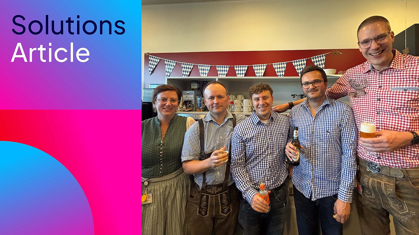 Fünf Personen stehen in Dirndl, Lederhosen und karierten Hemden mit Getränken vor einer blau-weißen Wimpelkette; links steht „Solutions Article“.