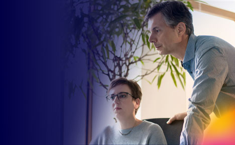 Junge Frau am Schreibtisch sitzend und mittelalter Mann neben ihr stehend, schauen auf einen PC.